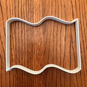 May include: A white plastic cookie cutter in the shape of a flag with a wavy top edge. The cutter is rectangular with rounded corners and sits on a wooden surface.