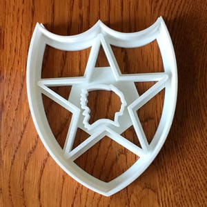 May include: A white plastic cookie cutter in the shape of a shield with a five-pointed star and a profile silhouette. The cutter is on a wooden surface.