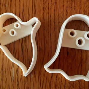 May include: Two white plastic ghost-shaped cookie cutters. Each cutter has a rectangular section with three circular holes for the eyes and mouth. The cutters are on a wooden surface.