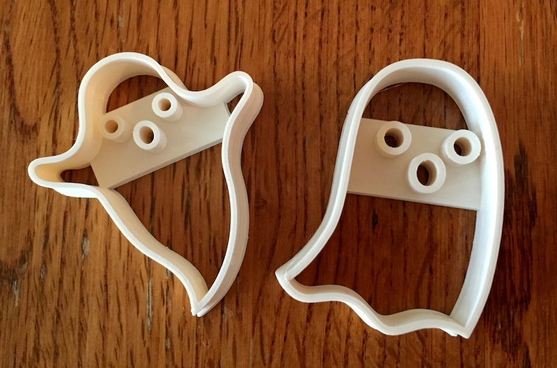 May include: Two white plastic ghost-shaped cookie cutters. Each cutter has a rectangular section with three circular holes for the eyes and mouth. The cutters are on a wooden surface.