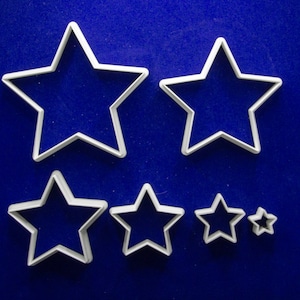 May include: A collection of white star-shaped cookie cutters in various sizes, arranged on a deep blue background. The cutters are made of a light-colored material and are designed with a simple, clean outline.