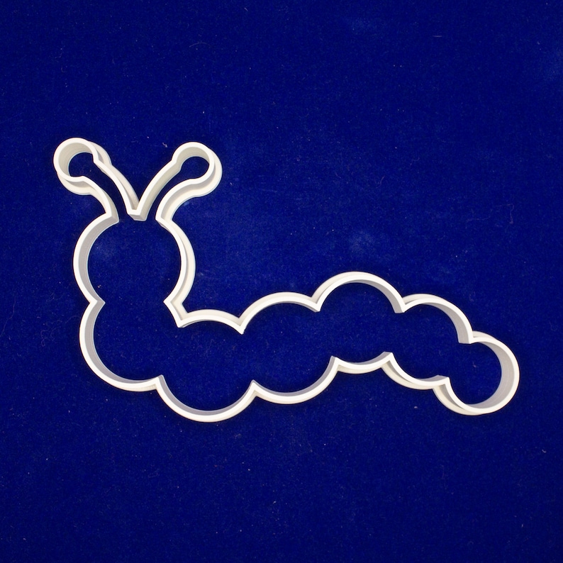 May include: A white plastic caterpillar-shaped cookie cutter. The cutter has a rounded head with antennae and a segmented body. The cutter is on a dark blue background.