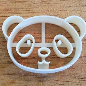 May include: A white plastic panda bear cookie cutter. The cutter is in the shape of a panda's head, with rounded ears, oval eyes, and a small nose and mouth. The cutter is designed to cut out panda-shaped cookies or other treats.