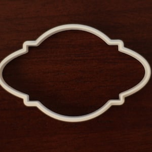 May include: A white plastic cookie cutter in an oval shape with decorative edges. The cutter is set against a dark brown wooden surface. The design is a classic shape for creating cookies or other baked goods.