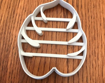 May include: A white plastic cookie cutter in the shape of a stylized flower or leaf. The cutter has a rounded outline with curved details and horizontal bars across the center. The cutter is on a wooden surface.