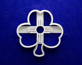 May include: A white plastic cookie cutter in the shape of a shamrock with a New Mexico Zia symbol in the center. The cookie cutter is on a blue background. The Zia symbol is a circle with four groups of four rays extending outward.