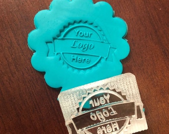 Puede incluir: Un sello turquesa con borde festoneado con el texto "Your Logo Here" en un diseño circular. Un sello rectangular transparente con el mismo diseño está al lado. Los sellos están sobre una superficie marrón oscuro.