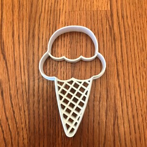 May include: A white plastic ice cream cone cookie cutter. The cutter features a scoop of ice cream with a scalloped edge and a waffle cone design. The cutter is placed on a wooden surface.