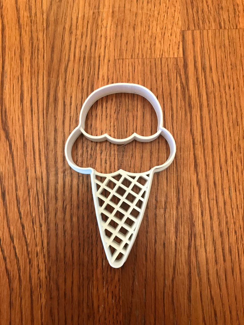 May include: A white plastic ice cream cone cookie cutter. The cutter features a scoop of ice cream with a scalloped edge and a waffle cone design. The cutter is placed on a wooden surface.