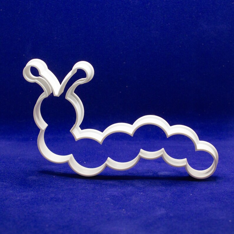 May include: A white plastic caterpillar-shaped cookie cutter. The cutter has a rounded head with two antennae and a segmented body. The design is simple and clean, perfect for creating fun-shaped cookies or fondant decorations.
