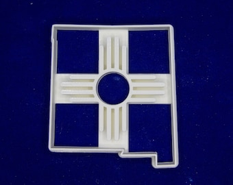 May include: A white, 3D-printed cookie cutter in the shape of the state of New Mexico. The design includes the Zia sun symbol in the center and four square cutouts. The cookie cutter is displayed against a dark blue background.