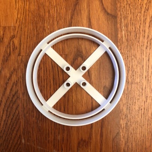 May include: A white plastic cookie cutter in a circular shape with an X design in the center. The cutter has a double-layered outer ring and four small holes where the X intersects. The cutter is on a wooden surface.