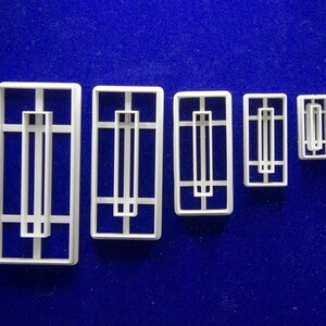 May include: A set of six white plastic cookie cutters in various sizes, arranged from largest to smallest. Each cutter has a rectangular frame with a central vertical bar and horizontal accents, set against a blue background. The design is reminiscent of a window or door.