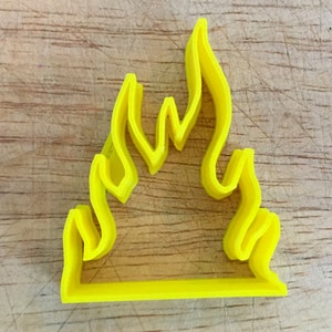 May include: A bright yellow flame-shaped cookie cutter. The cutter is made of plastic and has a simple, clean design. The flame shape is composed of several curved lines, with a flat base. The cutter is likely used for baking or crafting.
