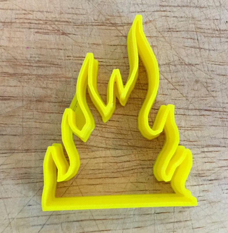 May include: A bright yellow flame-shaped cookie cutter. The cutter is made of plastic and has a simple, clean design. The flame shape is composed of several curved lines, with a flat base. The cutter is likely used for baking or crafting.