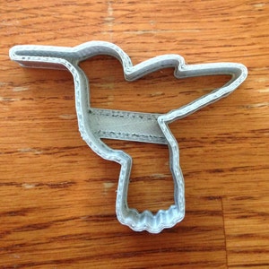 Puede incluir: Un cortador de galletas de pl&aacute;stico gris con forma de colibr&iacute;. El cortador tiene la forma de un colibr&iacute; con las alas extendidas. El cortador est&aacute; hecho de pl&aacute;stico en capas y se encuentra sobre una superficie de madera.