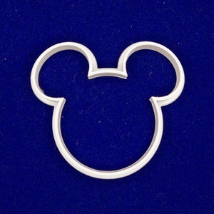 May include: A silver-colored cookie cutter in the shape of Mickey Mouse's head. The cutter has a rounded outline with two circular ears at the top and a curved bottom. The cutter is set against a dark blue background.