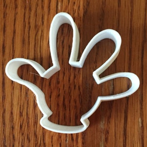 Puede incluir: Un cortador de galletas blanco de plástico con forma de mano. El cortador tiene un contorno detallado de una mano con los dedos extendidos. El diseño es adecuado para crear galletas con forma de mano u otros productos horneados. El cortador está colocado sobre una superficie de madera.