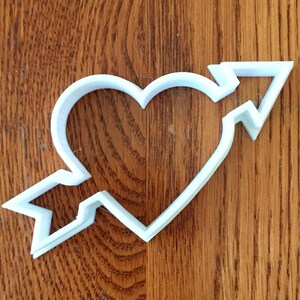 May include: A white plastic cookie cutter in the shape of a heart pierced by an arrow. The arrow's fletching and arrowhead are visible, and the heart shape is clearly defined. The cutter is placed on a wooden surface.