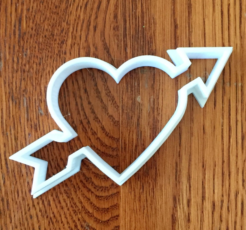 May include: A white plastic cookie cutter in the shape of a heart pierced by an arrow. The arrow's fletching and arrowhead are visible, and the heart shape is clearly defined. The cutter is placed on a wooden surface.