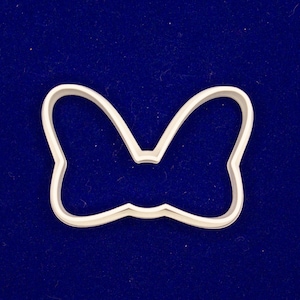 May include: A white plastic bow-shaped cookie cutter. The cutter is a solid outline of a bow, with rounded edges and a small indentation in the center. The cutter is set against a dark blue background.
