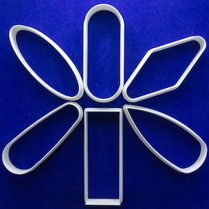 May include: White plastic cookie cutters in various geometric shapes, including ovals, rectangles, and a diamond, arranged in a flower-like pattern against a blue background. These cutters are likely used for baking and creating decorative cookies.
