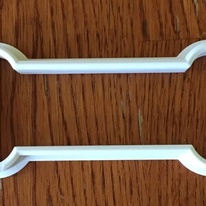 May include: A white plastic dog bone-shaped cookie cutter. The cutter has rounded ends and a rectangular center. The object is sitting on a wooden surface.