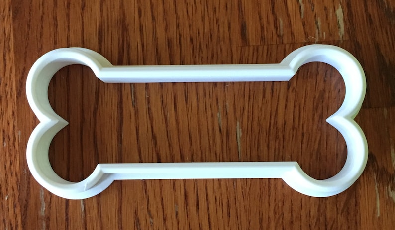May include: A white plastic dog bone-shaped cookie cutter. The cutter has rounded ends and a rectangular center. The object is sitting on a wooden surface.