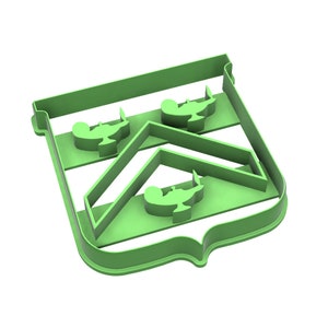 May include: A green plastic cookie cutter in the shape of a shield with three genie lamps. The cookie cutter has a decorative edge and a geometric interior design. The lamps are evenly spaced within the shield.