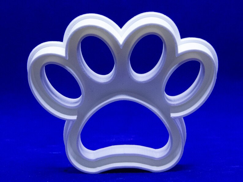 May include: A white, three-dimensional paw print cookie cutter. The cutter is made of a light-colored plastic and has a layered design. The background is a solid, deep blue.