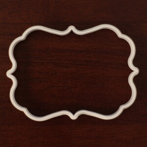 May include: A white, ornate-shaped cookie cutter. The cutter has a decorative, scalloped edge and is set against a dark brown wooden surface. The shape is a rectangle with curved sides.