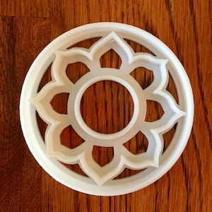May include: A white plastic cookie cutter in the shape of a flower with a circular center and petal-like shapes around the edge. The cutter is sitting on a wooden surface.