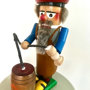 May include: Wooden nutcracker figurine of a man with a red hat, blue sleeves, and brown overalls. The figurine has a white beard, a red nose, and is holding a barrel. The base is green with yellow accents.