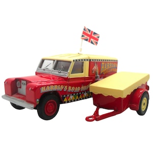 Puede incluir: Un Land Rover de juguete rojo con techo amarillo y blanco y un remolque. El Land Rover tiene "HARRIS'S ROAD SHOW" escrito en el lateral y una bandera de la Unión Jack en la parte superior. El remolque tiene una lona amarilla y "HARRIS'S GALLOPERS" escrito en el lateral.