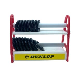 Puede incluir: Un estante rojo en miniatura para neum&aacute;ticos con dos estantes blancos que sostienen pilas de neum&aacute;ticos negros. El estante tiene una base amarilla con el logotipo de Dunlop. Los neum&aacute;ticos son peque&ntilde;os, probablemente para un coche de juguete.