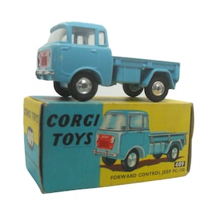 May include: A blue toy truck with a white cab and a flatbed. The truck is on a yellow box with the words "Corgi Toys" and "Forward Control Jeep FC-150" printed on it. The box also has the number "409" printed on it.