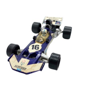 May include: A blue and white toy race car with the number 16 on the side. The car has a driver in the cockpit and the words "Surtees" on the front.