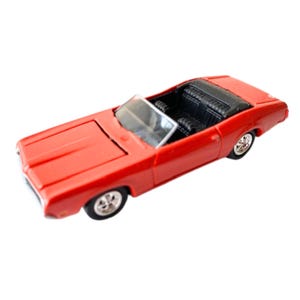 May include: A red die-cast convertible toy car featuring a black interior, a clear plastic windscreen, and silver-coloured wheels with black tyres. The vehicle has a long bonnet, a flat boot, and a detailed dashboard visible through the open top.