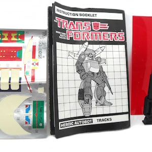 Op de afbeelding: Een instructieboekje voor het Transformers-speelgoed Tracks. Het boekje bevat een zwart-witte illustratie van Tracks in robotmodus. Het boekje bevat ook een vel met stickers met verschillende stickers voor het speelgoed. De tekst op het boekje luidt "The Transformers" en "Heroic Autobot Tracks".