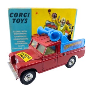 May include: A vintage Corgi Toys Land Rover truck toy. The red truck features blue loudspeakers, a clown figure, and a yellow sign that reads "The Circus Is Here." The box behind the truck has the Corgi Toys logo.