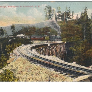 Puede incluir: Postal vintage que muestra un tren cruzando el puente "S" en Alamogordo a Cloudcroft, N.M. La imagen muestra un tren de vapor sobre un puente de madera curvado, rodeado de árboles verdes y un paisaje rocoso. El cielo es azul pálido.
