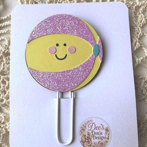 May include: A handmade paper clip featuring a smiling yellow face with pink cheeks and a purple glittery band. The paper clip is attached to a white card with the text "Dee's Doxie Designs" and a floral illustration.
