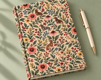 Könnte beinhalten: Ein rechteckiges Notizbuch mit cremefarbenem Einband, das ein wiederkehrendes Muster aus verschiedenen Dackeln zwischen roten, gelben und blauen Blumen mit grünem Blattwerk zeigt. Ein eleganter, heller Kugelschreiber liegt daneben.