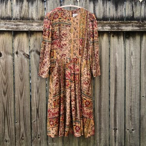 May include: A long-sleeved midi dress featuring a multi-panel patchwork design with a repeating floral and geometric pattern in shades of deep red, mustard yellow, olive green, and cream. The dress has a round neckline and a pleated skirt.