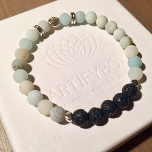 以下が含まれることがあります： 水色、クリーム色、黒色のビーズを組み合わせたブレスレット。シルバーカラーのアクセントがあり、「ARTIFYBOX INSPIRATIONAL GEMSTONE JEWELRY」の文字が入った白い箱に入っています。