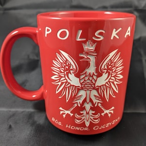 May include: A red ceramic mug with a white Polish eagle emblem and the word "POLSKA" in white lettering. The mug also features the text "BÓG, HONOR, OJCZYZNA". The mug has a curved handle.