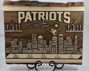 May include: A wooden sign with a brown background and white text. The sign features the New England Patriots logo and the text "PATRIOTS" in large letters. The sign also includes the text "2018 CHAMPIONS" and a list of players' names.