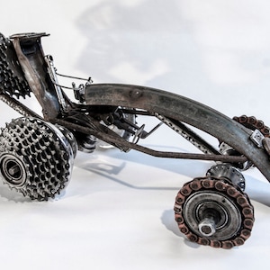May include: A sculpture of a race car made from recycled bicycle parts. The car is made of metal and has a rusty patina. The wheels are made from bicycle gears and chains.