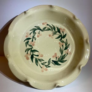 May include: A round ceramic pie dish with a cream-colored glaze and a scalloped edge. The interior features a hand-painted wreath design consisting of dark green leaves and small light pink flowers, accented by tiny white dots arranged in a circle.