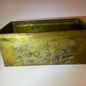 May include: A rectangular brass planter with a weathered, golden-brown patina. The front panel features an etched, light-colored design depicting two deer standing in a landscape with stylized pine trees and grassy ground details.